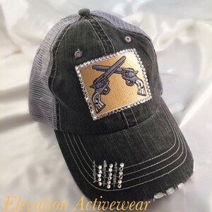 May include: A black and gray distressed trucker hat with a gold patch featuring two crossed pistols with rhinestones around the border. The hat has a gray mesh back and rhinestones on the bill.