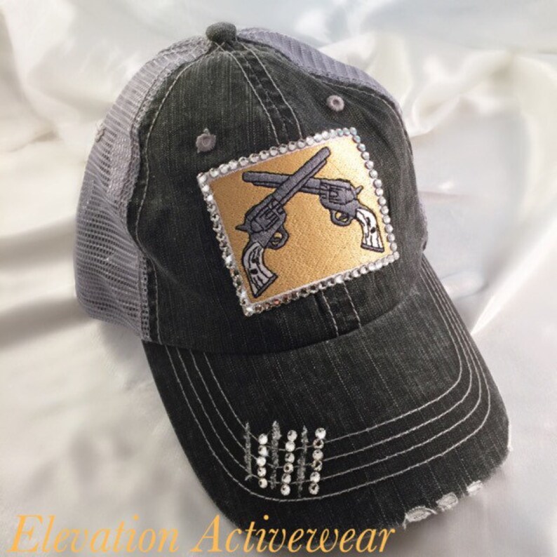 May include: A black and gray distressed trucker hat with a gold patch featuring two crossed pistols with rhinestones around the border. The hat has a gray mesh back and rhinestones on the bill.