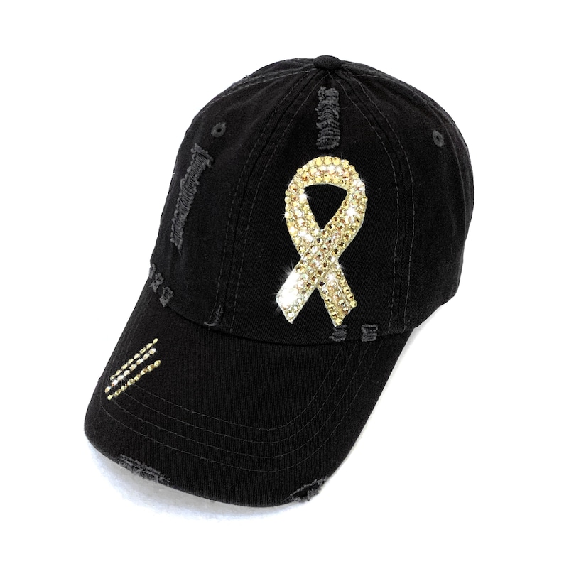 May include: Black distressed baseball cap with a sparkling gold ribbon design on the front. The cap has a curved brim and decorative rhinestone accents. The cap is a stylish accessory.