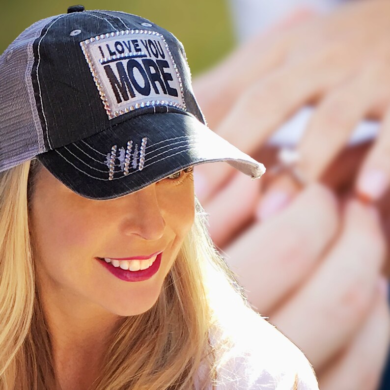 K&ouml;nnte beinhalten: Dunkelblaue Jeans-Baseballkappe mit grauem Mesh-R&uuml;cken. Auf der Kappe steht in Wei&szlig; und Silber der Text "I LOVE YOU MORE". Strasssteine ​​sind ebenfalls auf der Kappe vorhanden.