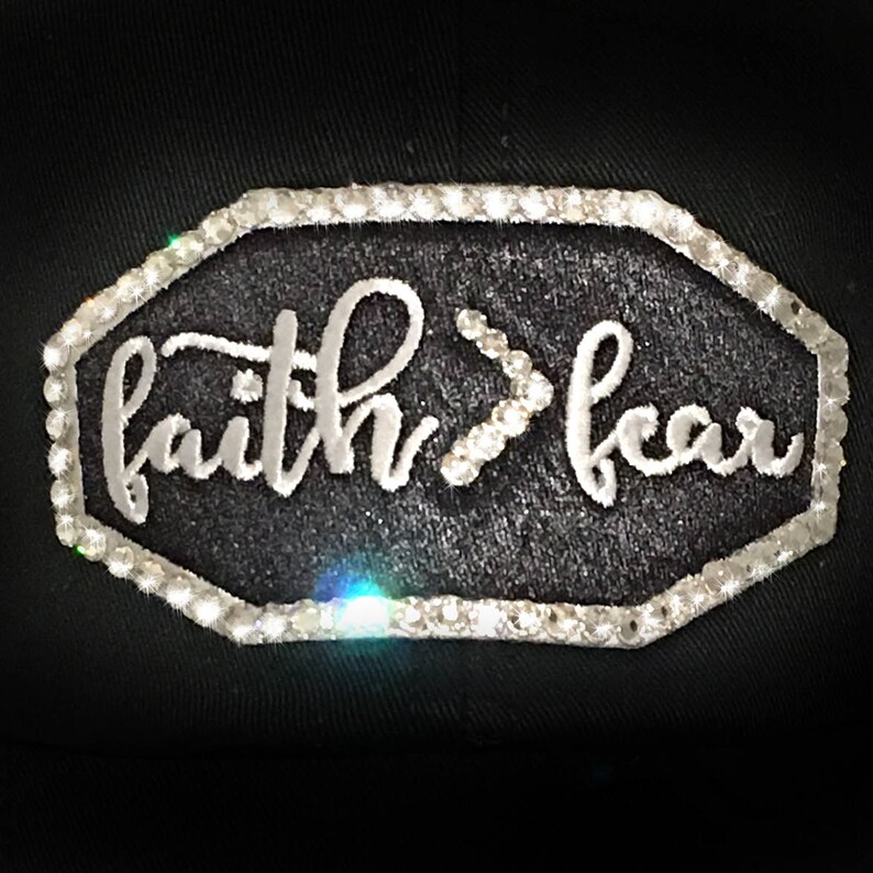 Puede incluir: Gorra de b&eacute;isbol negra con un parche bordado blanco que dice "faith>fear" rodeado de diamantes de imitaci&oacute;n.