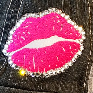 May include: A pink embroidered lip design with a clear rhinestone border. The lip design is on a dark denim background.