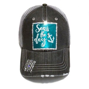 Puede incluir: Una gorra de camionero gris y blanca con un parche cuadrado turquesa que dice "Seas the day" con un ancla blanca. El parche está adornado con diamantes de imitación.