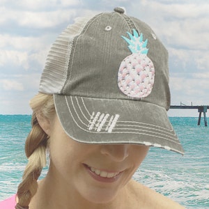 May include: A distressed gray trucker hat with a white mesh back. The front of the hat features a bedazzled pineapple design with pink gems and a turquoise top. The hat has a curved brim and a distressed look.