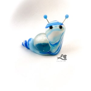 May include: A blue and white glass snail sculpture with black eyes and blue antennae. The snail is curled up in a spiral shape.