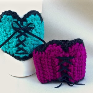 May include: Two crocheted cup cozies in the shape of a corset. The larger one is teal with black ribbon lacing and a black bow. The smaller one is magenta with black ribbon lacing and a black bow.