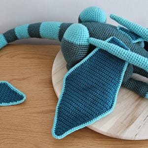May include: A handmade crochet dragon in shades of teal, blue, and gray. The dragon features a long tail with a fin, large wings, and a detailed head with horns. The plush toy is resting on a wooden surface.