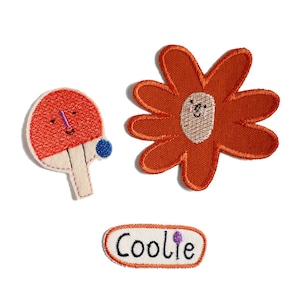 Op de afbeelding: Drie geborduurde strijkpatches: een bloem met een glimlachend gezicht, een pingpongbat met een glimlachend gezicht en een wit ovaal patch met het woord "Coolie" in zwarte letters.