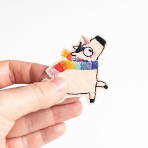 Op de afbeelding: Een crème-kleurige eenhoorn patch met een regenboog sjaal. De patch heeft zwarte geborduurde details voor de ogen, benen en hoorn. De sjaal is een levendige reeks kleuren. De patch wordt in de hand van een persoon gehouden.