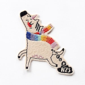 Könnte beinhalten: Gestickter Aufnäher mit einem skurrilen Einhorn mit Regenbogen-Schal. Das Einhorn ist cremefarben mit schwarzen Akzenten, einem rosa Horn und einer schwarzen Mähne. Der Aufnäher enthält die Buchstaben "NZS" in einem schwarzen Rechteck.