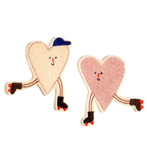Op de afbeelding: Twee geborduurde vilt harten op een witte achtergrond. Beide harten hebben bruine benen en dragen rolstoeltjes. Een hart is beige en heeft een blauwe hoed. Het andere hart is roze.