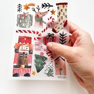 Könnte beinhalten: Eine Blatt mit transparenten Aufklebern mit einem Aquarell-Weihnachtsmotiv. Die Aufkleber zeigen einen Nussknacker, ein Rentier, einen Strumpf, ein Haus, Zuckerstangen, Geschenke und Weihnachtsbäume. Die Aufkleber sind perfekt, um Ihren Weihnachtsbastelarbeiten eine festliche Note zu verleihen.