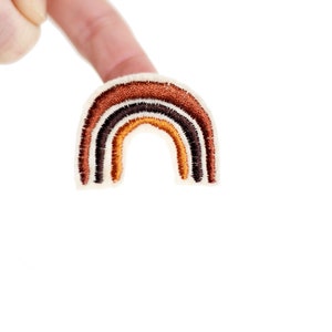 Può includere: Una piccola toppa ricamata a forma di arcobaleno con strisce marroni, arancioni e nere su uno sfondo bianco.