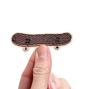 Könnte beinhalten: Ein kleiner gestickter Aufnäher eines braunen Skateboards mit weißen Rädern. Der Aufnäher wird von einem Finger gehalten.