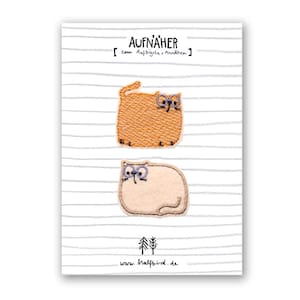 May include: A white card with two embroidered cat patches. The top cat is orange with glasses, and the bottom cat is beige. The card has gray horizontal stripes and the text "AUFNÄHER" and "www.halfbird.de".
