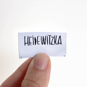 Puede incluir: Una etiqueta tejida blanca con bordado negro que dice "HEIDEWITZKA".
