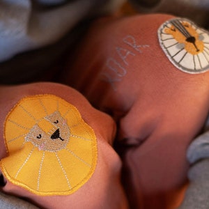 Może przedstawiać: Różowe spodnie dresowe z haftowanym żółtym lewią głową na jednej nogawce i haftowanym napisem "Roar" na drugiej nogawce.