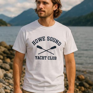 May include: White t-shirt with the text "HOWE SOUND YACHT CLUB" in navy blue. The design includes crossed oars. The person is wearing khaki pants and standing on a rocky shoreline with a lake and mountains in the background.