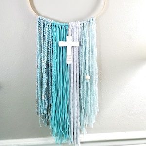 May include: A handcrafted wall hanging featuring a wooden hoop and cascading strands of blue and white yarn. A white cross with the words "Bless Your Heart" hangs in the center. The piece is designed for wall decor.