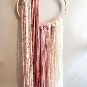 May include: A handmade wall hanging featuring a wooden hoop with strands of pink, cream, and white yarn. The yarn is arranged in vertical lines, creating a textured and layered effect. The wall decor is designed to add a touch of bohemian style to any room.
