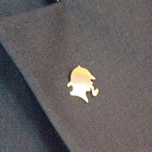 Può includere: Una piccola spilla da bavero in metallo dorato a forma di silhouette di una persona che indossa un cappello deerstalker e fuma una pipa. La spilla è fissata sul tessuto blu scuro di un blazer e presenta una superficie metallica liscia e lucida.