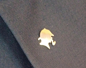 May include: A small, gold-toned metal lapel pin shaped like a silhouette of a person wearing a deerstalker hat and smoking a pipe. The pin is attached to the dark navy blue fabric of a blazer, showing a smooth, reflective metallic surface.