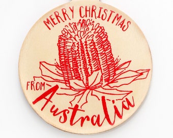 May include: A round wooden ornament featuring a red line drawing of a banksia flower. The text "MERRY CHRISTMAS FROM AUSTRALIA" is printed in red ink. The ornament includes a small wooden loop at the top with a piece of brown twine attached.