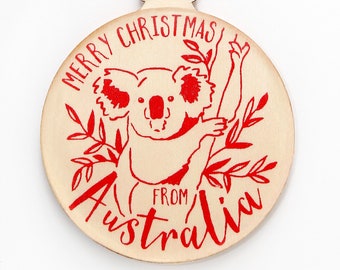 May include: A round wooden ornament featuring a red graphic of a koala in a tree with the text MERRY CHRISTMAS FROM AUSTRALIA printed in red. The ornament has a natural light wood finish and is suspended by a thin brown twine loop.