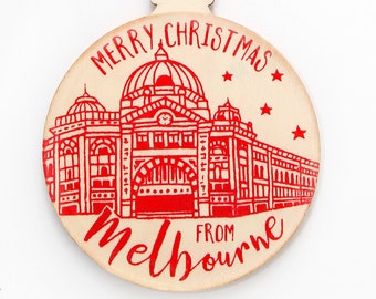 May include: A round wooden Christmas ornament featuring a red ink illustration of Flinders Street Station in Melbourne. The text reads MERRY CHRISTMAS and FROM MELBOURNE in red script. The ornament hangs from a natural jute twine loop.