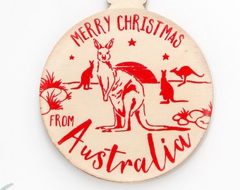 May include: A round wooden Christmas ornament featuring a red printed design of kangaroos in a landscape with the text MERRY CHRISTMAS FROM AUSTRALIA. The ornament has a light wood grain finish and is attached to a piece of natural jute twine.