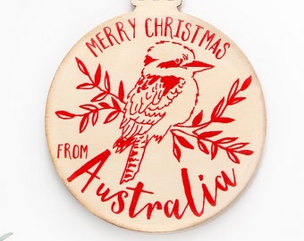 May include: A round wooden ornament featuring a red graphic of a kookaburra perched on a branch. The text reads Merry Christmas from Australia in red ink. The ornament includes a jute twine loop for hanging and a natural light wood finish.