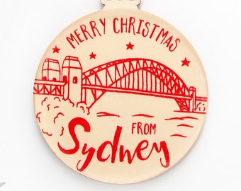 May include: A round, light-colored wooden Christmas ornament featuring a red line-art illustration of the Sydney Harbour Bridge. The text reads MERRY CHRISTMAS FROM SYDNEY in red ink. The ornament hangs from a natural jute twine loop.