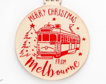 May include: A round, light-colored wooden ornament hanging from a jute twine loop. The ornament features a red graphic of a Melbourne tram and a Christmas tree, with the red text "MERRY CHRISTMAS FROM Melbourne" and small red stars printed on it.
