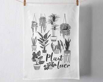 May include: A white rectangular kitchen towel featuring a black line art illustration of various potted houseplants and hanging planters. The text "Plant lover" is printed in a black script font in the center of the towel design.