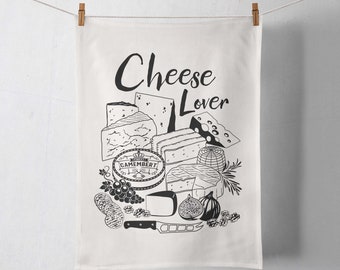 May include: A white kitchen towel featuring a black line art illustration of various cheeses, grapes, figs, walnuts, and a cheese knife. The text "Cheese Lover" is printed in black at the top. The towel is suspended from a string by two wooden clips.