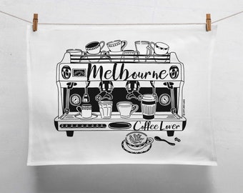 May include: A white tea towel featuring a black graphic illustration of an espresso machine with various coffee cups and the text Melbourne Coffee Lover. The design includes a latte art cup and a spoon below the machine, all printed in black ink.