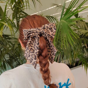 May include: A white t-shirt with a blue and red graphic print that says "New York Big City of Dreams 0000 90s united different music of the 90s Bronx Queens Manhattan". The person is wearing a brown and black leopard print hair bow.