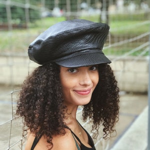May include: A woman with curly brown hair is wearing a black leather newsboy cap with a textured surface. She is wearing a black tank top.