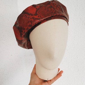 Puede incluir: Un gorro rojo con estampado de piel de serpiente. El gorro está en una cabeza de maniquí blanca.