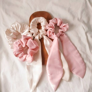 May include: Three satin hair scrunchies, one white and two pink. The white scrunchie has a long tail, and the pink scrunchies each have two tails.