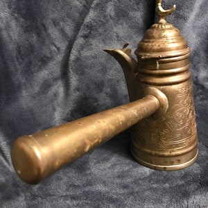 May include: A brass coffee pot with an ornate design and a bird finial on the lid. The pot has a long, curved handle and a spout on the side.