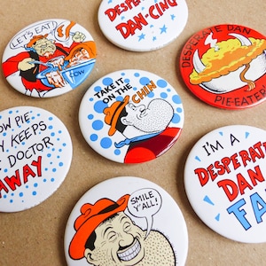 Pode incluir: Nove botões redondos e coloridos com várias imagens e textos. Os botões apresentam um personagem de desenho animado chamado Desperate Dan, um bolo de vaca e o texto "Um bolo de vaca por dia mantém o médico longe".