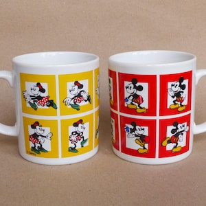 May include: Two white ceramic mugs with handles. One mug has a yellow background with images of Mickey Mouse and Minnie Mouse in red and black. The other mug has a red background with images of Mickey Mouse and Minnie Mouse in black and white. Both mugs have the text "Disney" on the bottom.