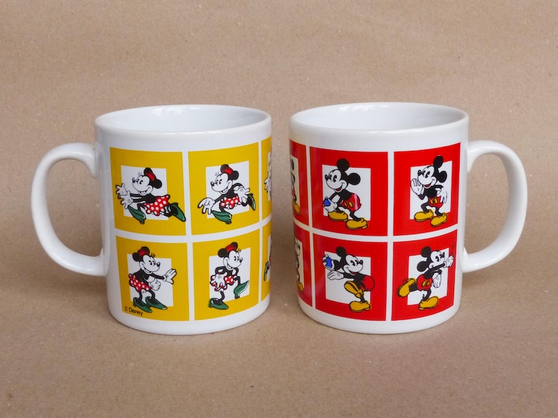 May include: Two white ceramic mugs with handles. One mug has a yellow background with images of Mickey Mouse and Minnie Mouse in red and black. The other mug has a red background with images of Mickey Mouse and Minnie Mouse in black and white. Both mugs have the text "Disney" on the bottom.