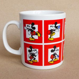 May include: White ceramic mug with a red and white design featuring Mickey Mouse in different poses. The mug has a handle and is decorated with a red background and white squares.