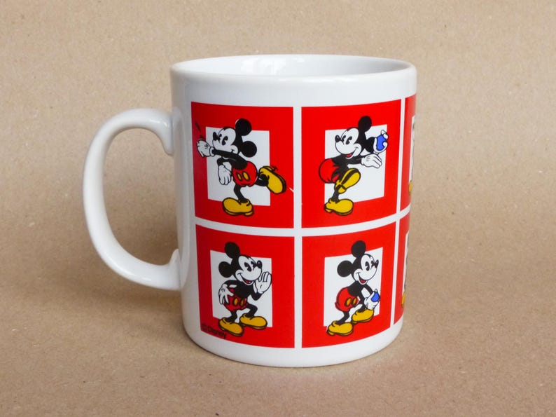 May include: White ceramic mug with a red and white design featuring Mickey Mouse in different poses. The mug has a handle and is decorated with a red background and white squares.
