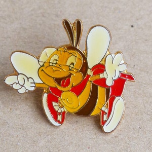 Op de afbeelding: Een gouden speld met een cartoon-bij die rode schoenen en een rode strik draagt. De bij glimlacht en heeft zijn tong uitgestoken. De bij zwaait met zijn rechterhand en wijst met zijn linkerhand.