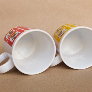 May include: Two white ceramic mugs with handles. One mug has a red and yellow design, the other has a yellow and orange design.
