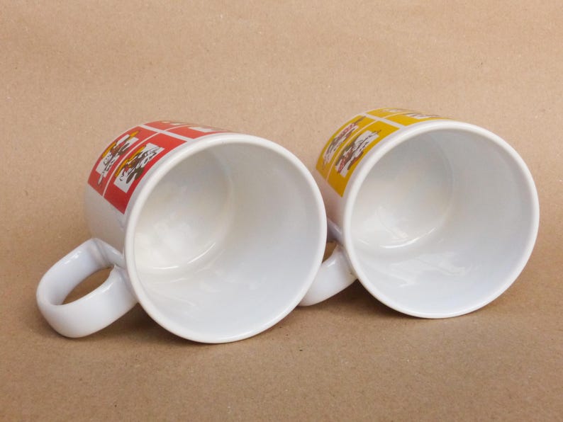 May include: Two white ceramic mugs with handles. One mug has a red and yellow design, the other has a yellow and orange design.