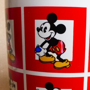 May include: White ceramic mug with a red and white pattern featuring multiple images of Mickey Mouse. The mug has a red border around each image of Mickey Mouse.